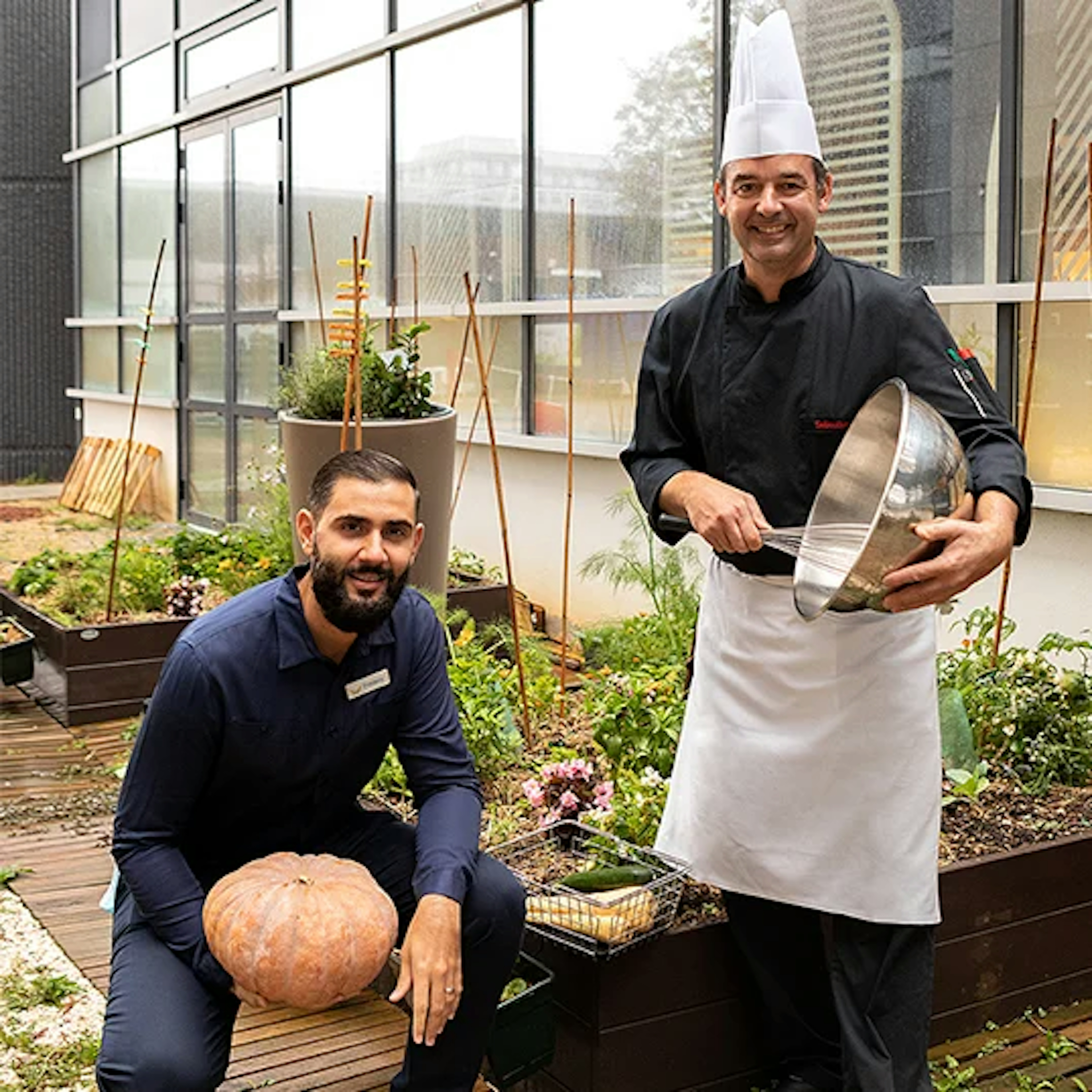 Two men, one holding a pumpkin and the other a whisk, stand smiling in an outdoor garden with raised plant beds.