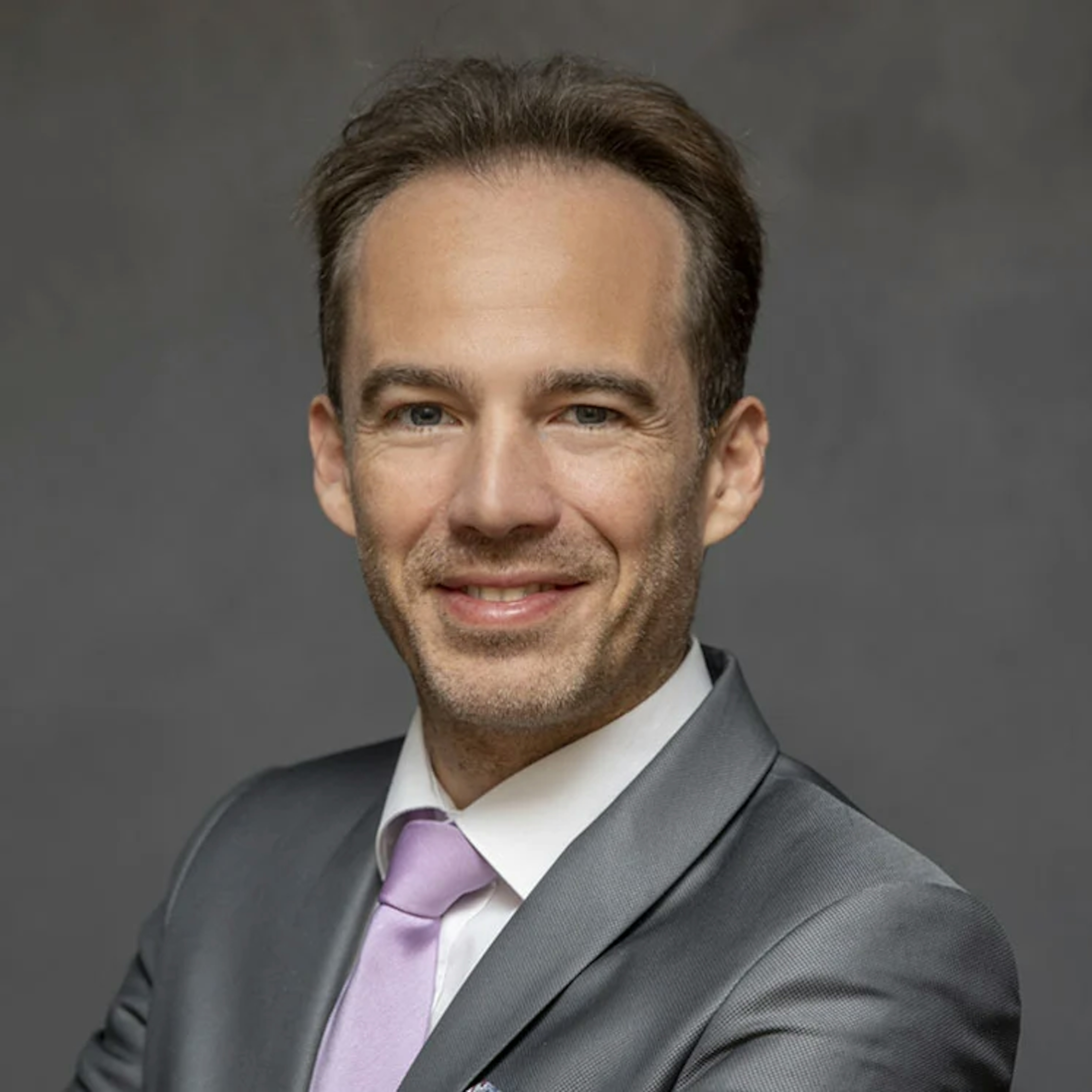 Portrait of Fabrice Carré, a man with a slight beard, wearing a light gray suit, white shirt, and purple tie, smiling against a gray background.