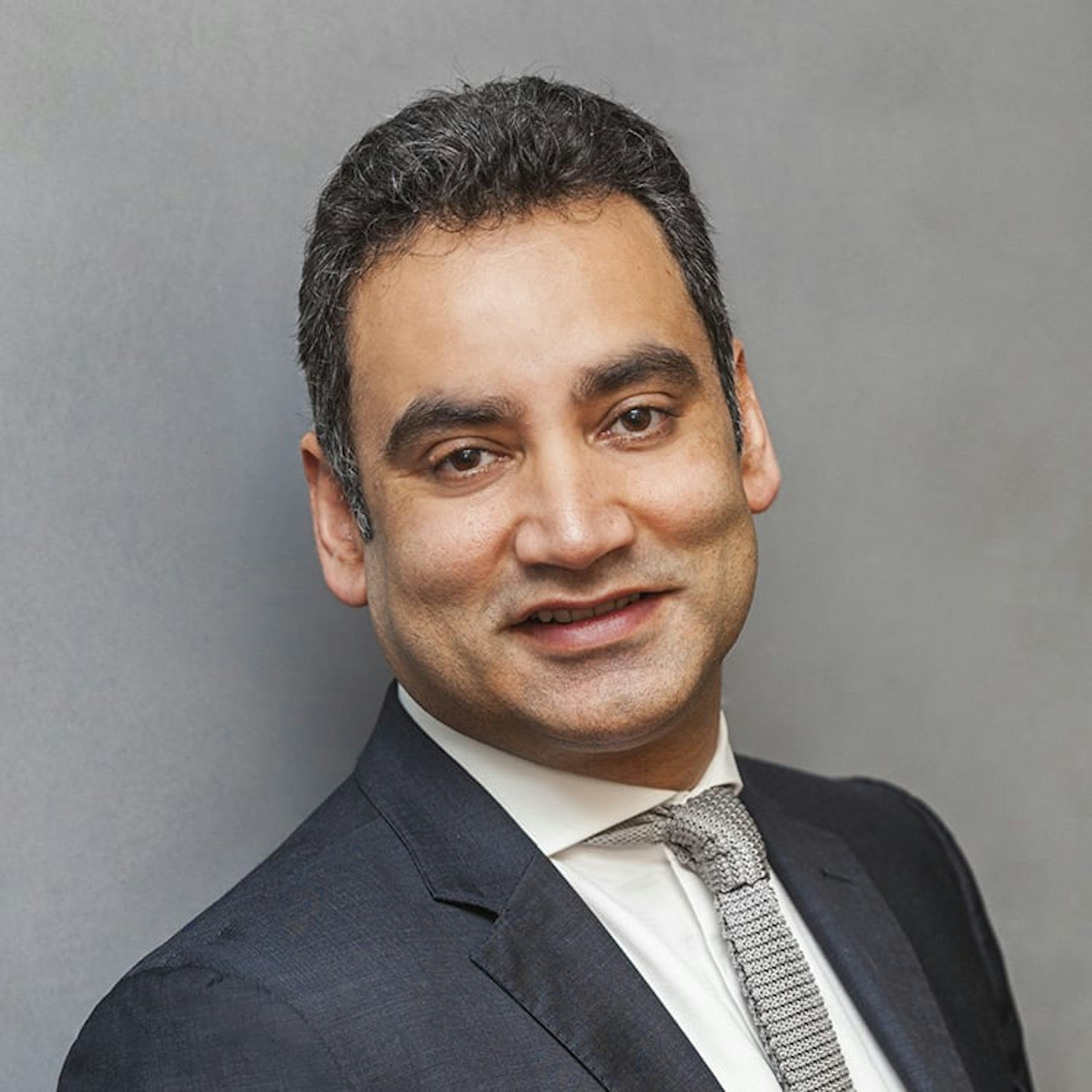 Portrait of Gaurav Bhushan, a man with short dark hair, wearing a dark suit, white shirt, and blue patterned tie, smiling.