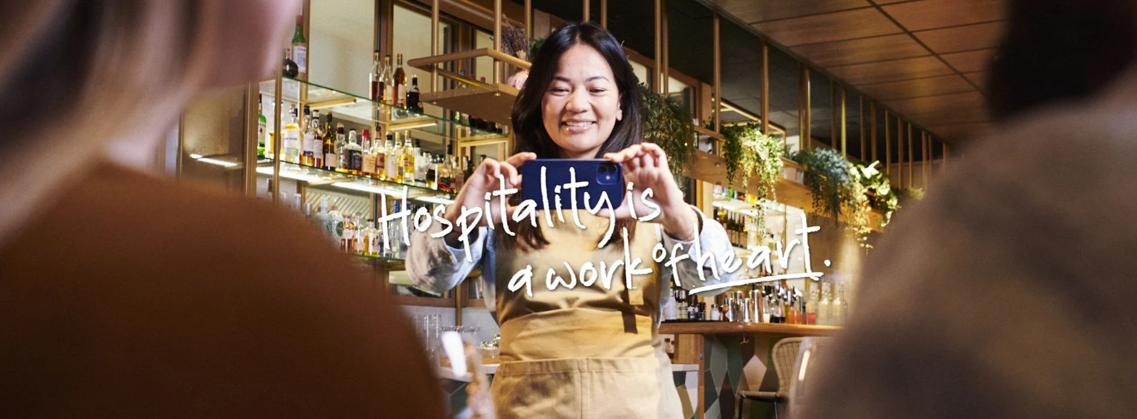 A woman taking a picture in a bar with the text: Hospitality is a work of heart