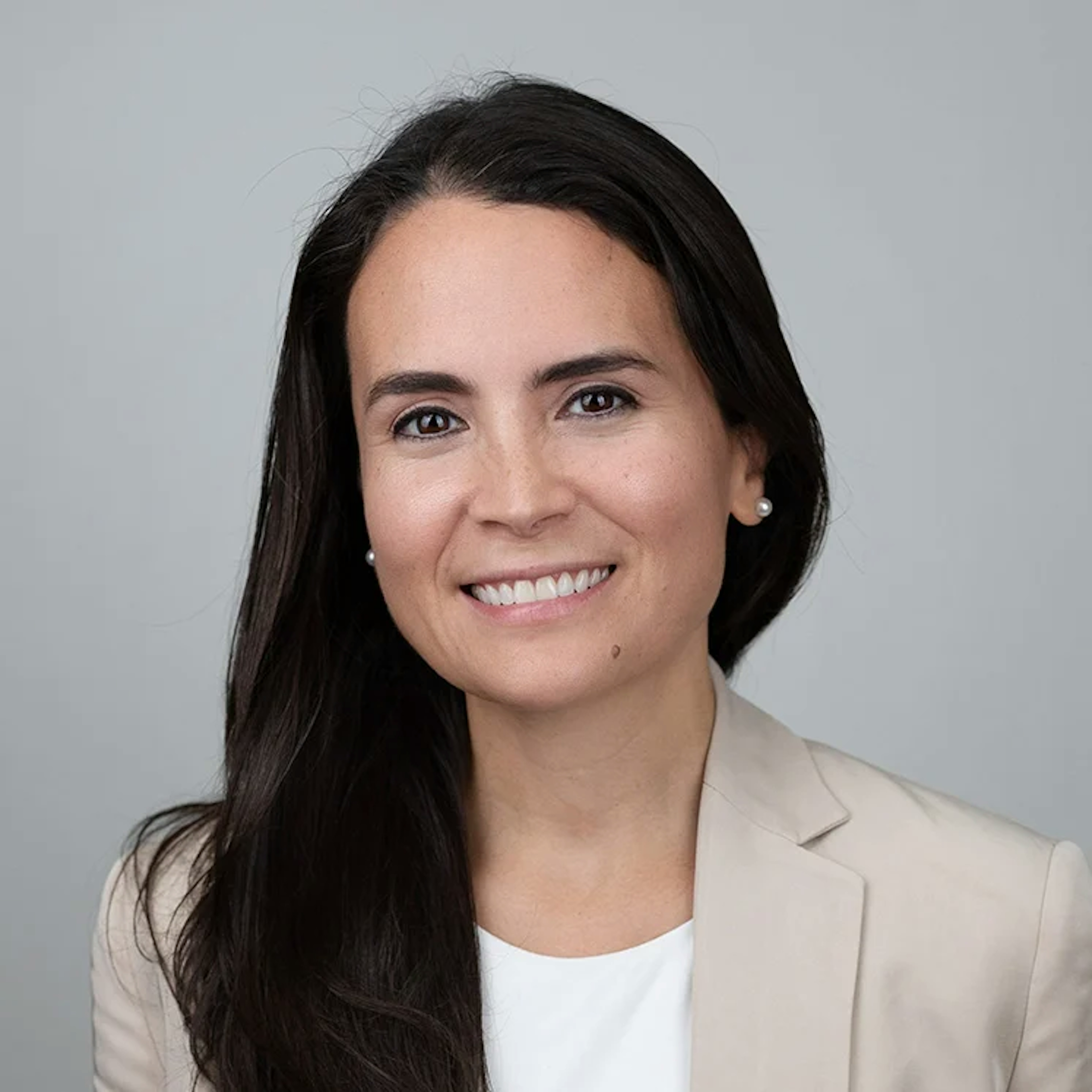 Portrait of Gilda Perez-Alvarado, a woman with long dark hair, wearing a white top and a beige blazer, smiling.