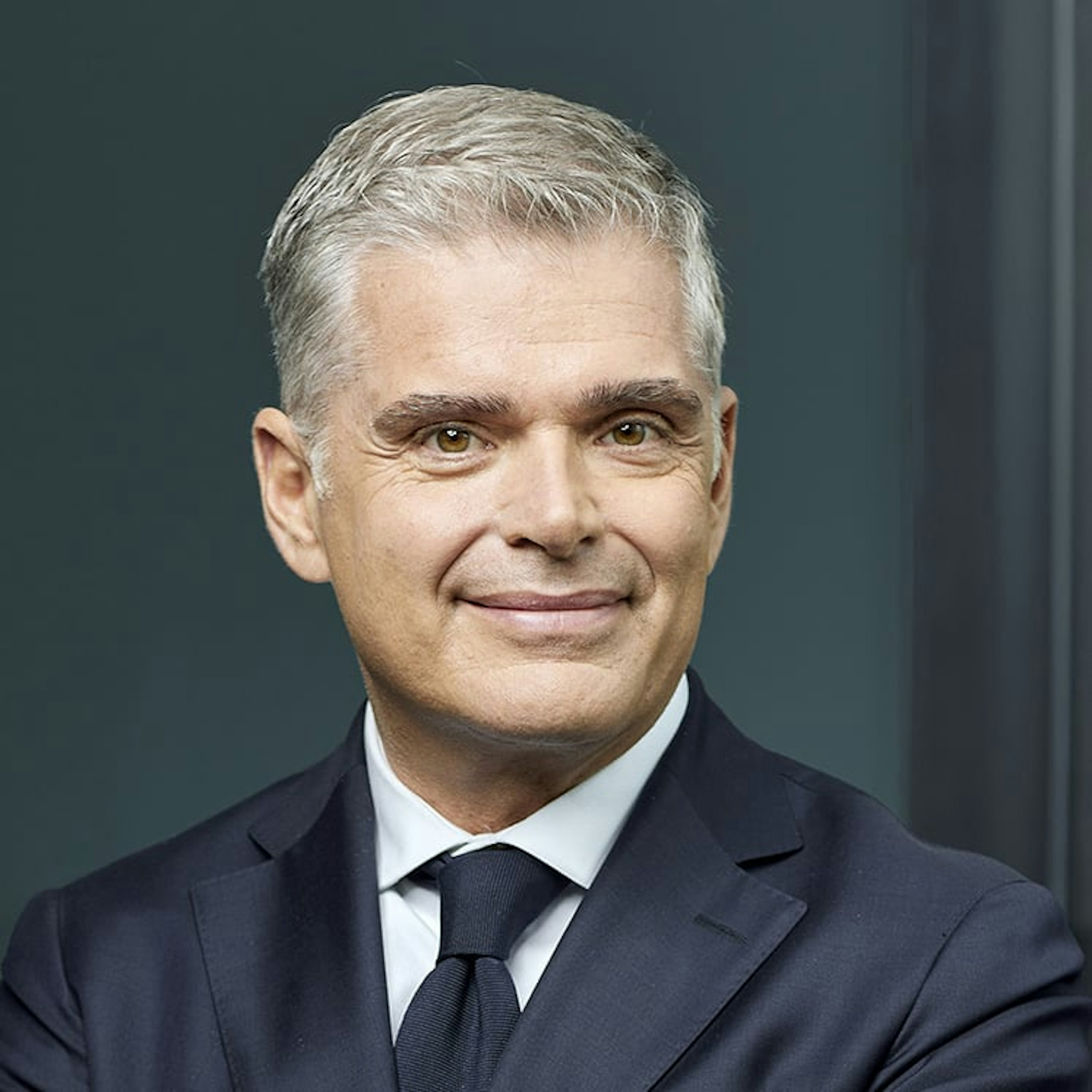 ortrait d'Omer Acar, un homme aux cheveux gris courts, portant un costume foncé, une chemise blanche et une cravate sombre, souriant sur fond discret.