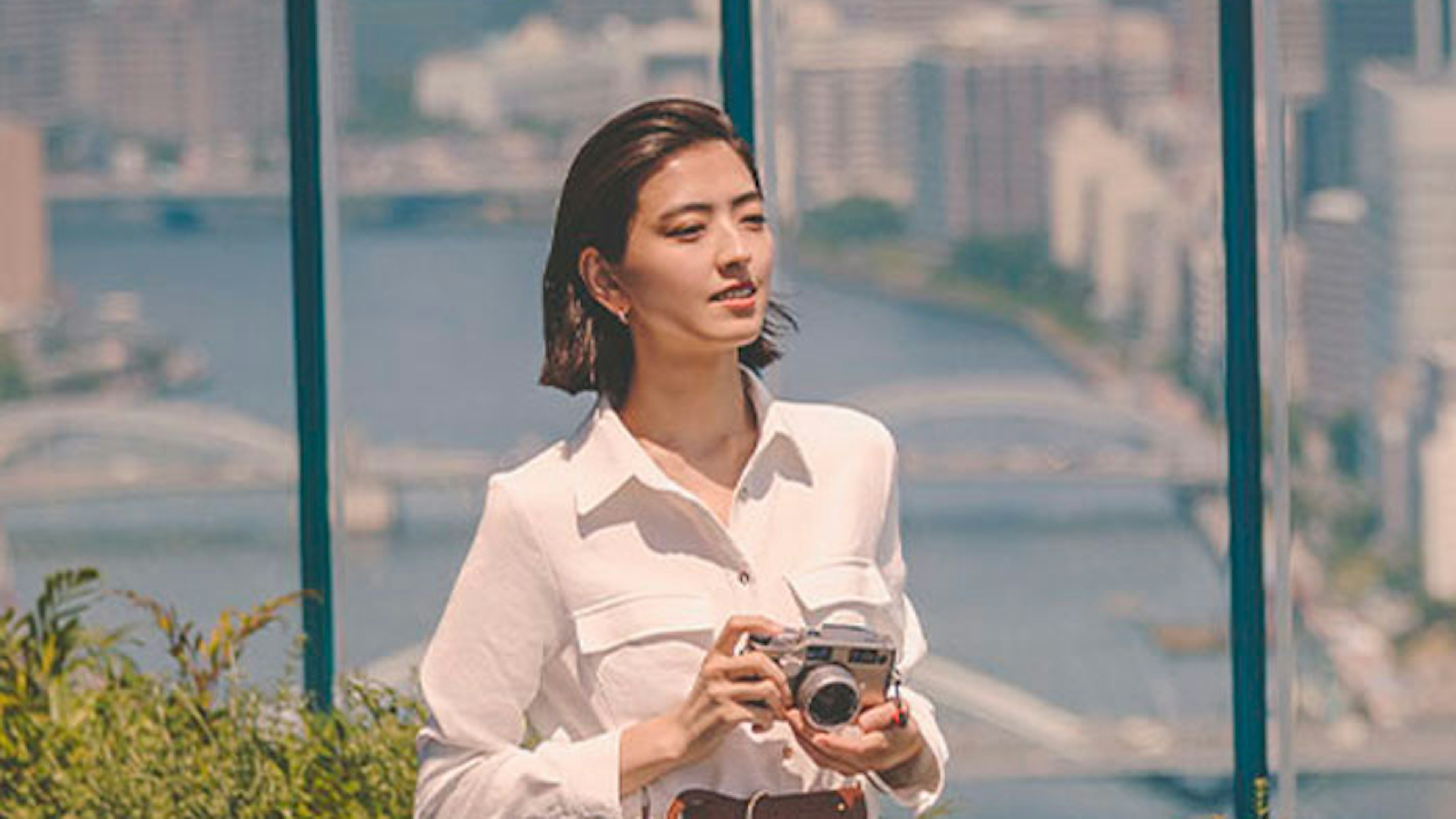 A woman in a white dress and brown belt holds a camera on a rooftop with a city skyline and water in the background.