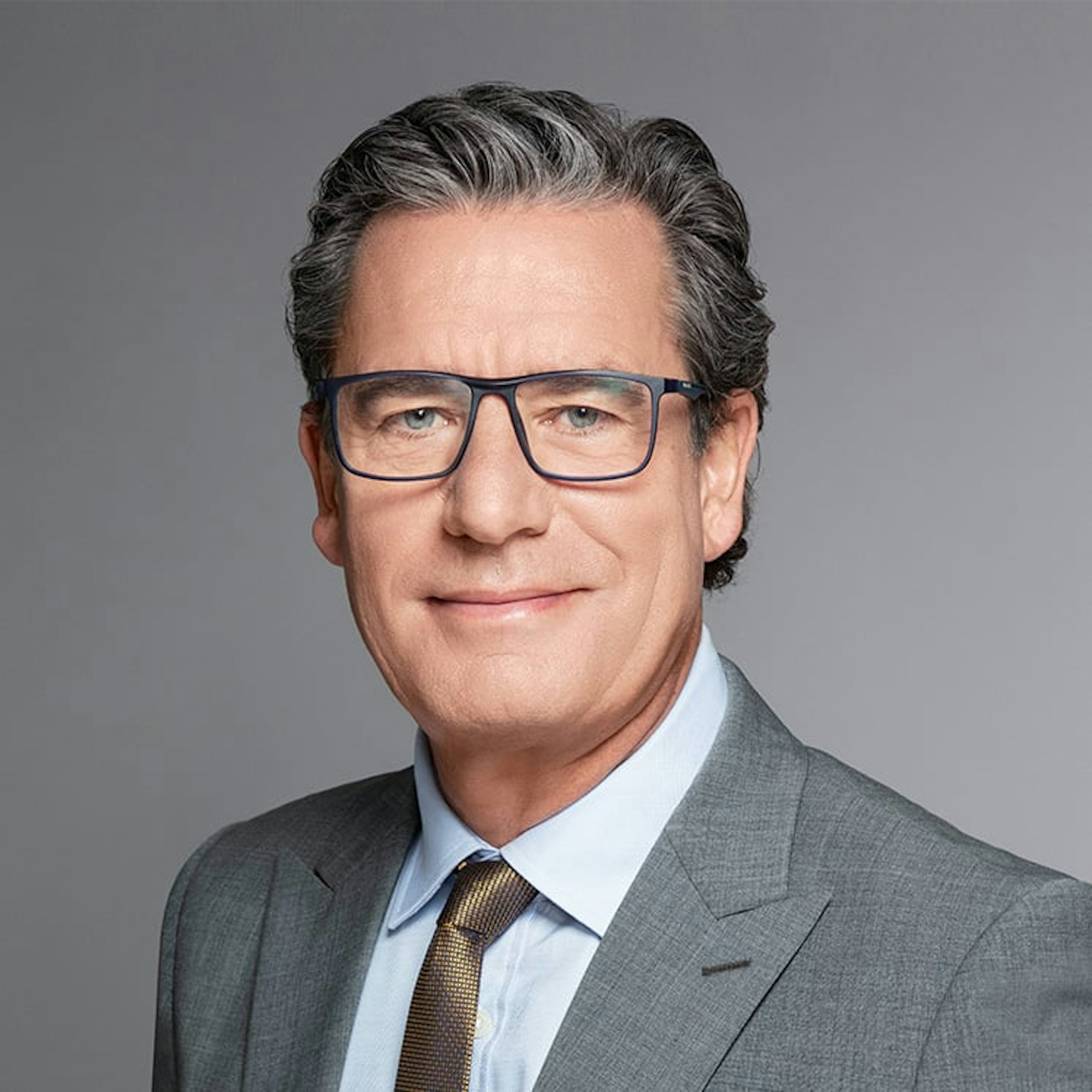 Portrait of Thomas Dubaere, a man with grey wavy hair, glasses, wearing a light grey suit and gold-patterned tie, smiling.