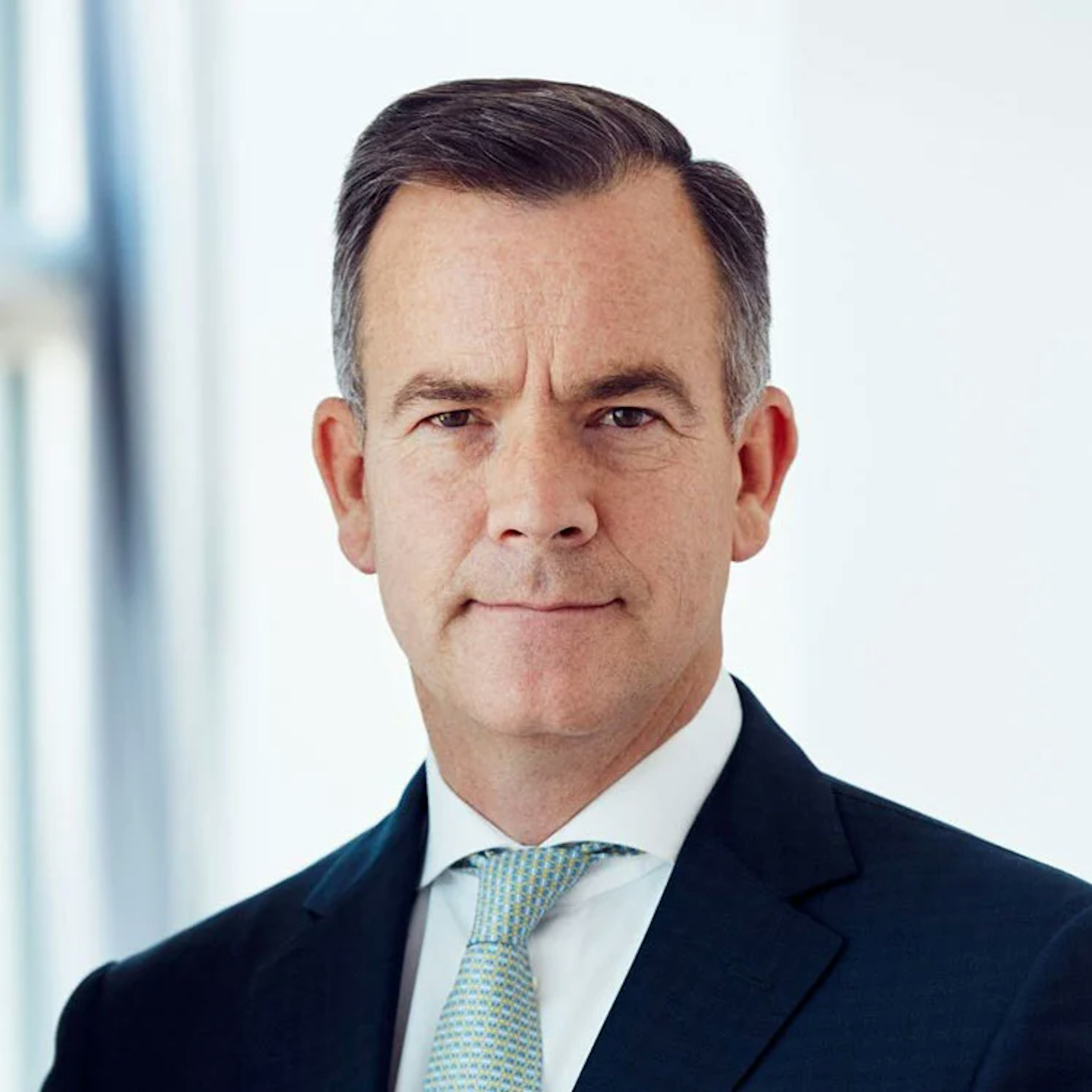 Portrait of Duncan O'Rourke, a man with neatly styled grey hair, wearing a dark suit, white shirt, and patterned tie, looking directly at the viewer.