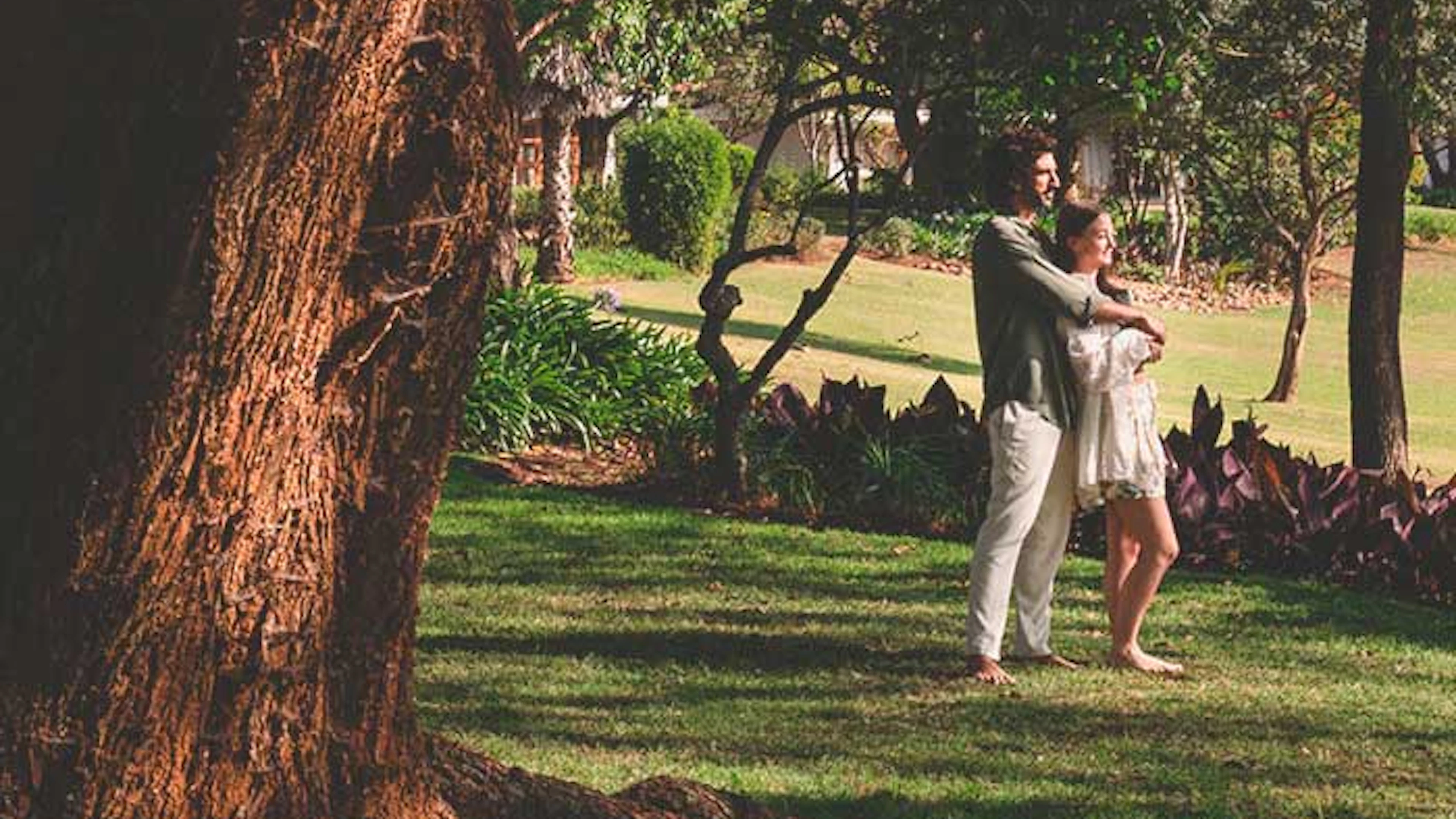 Un couple s'embrasse dans un jardin verdoyant et ensoleillé avec de grands arbres.