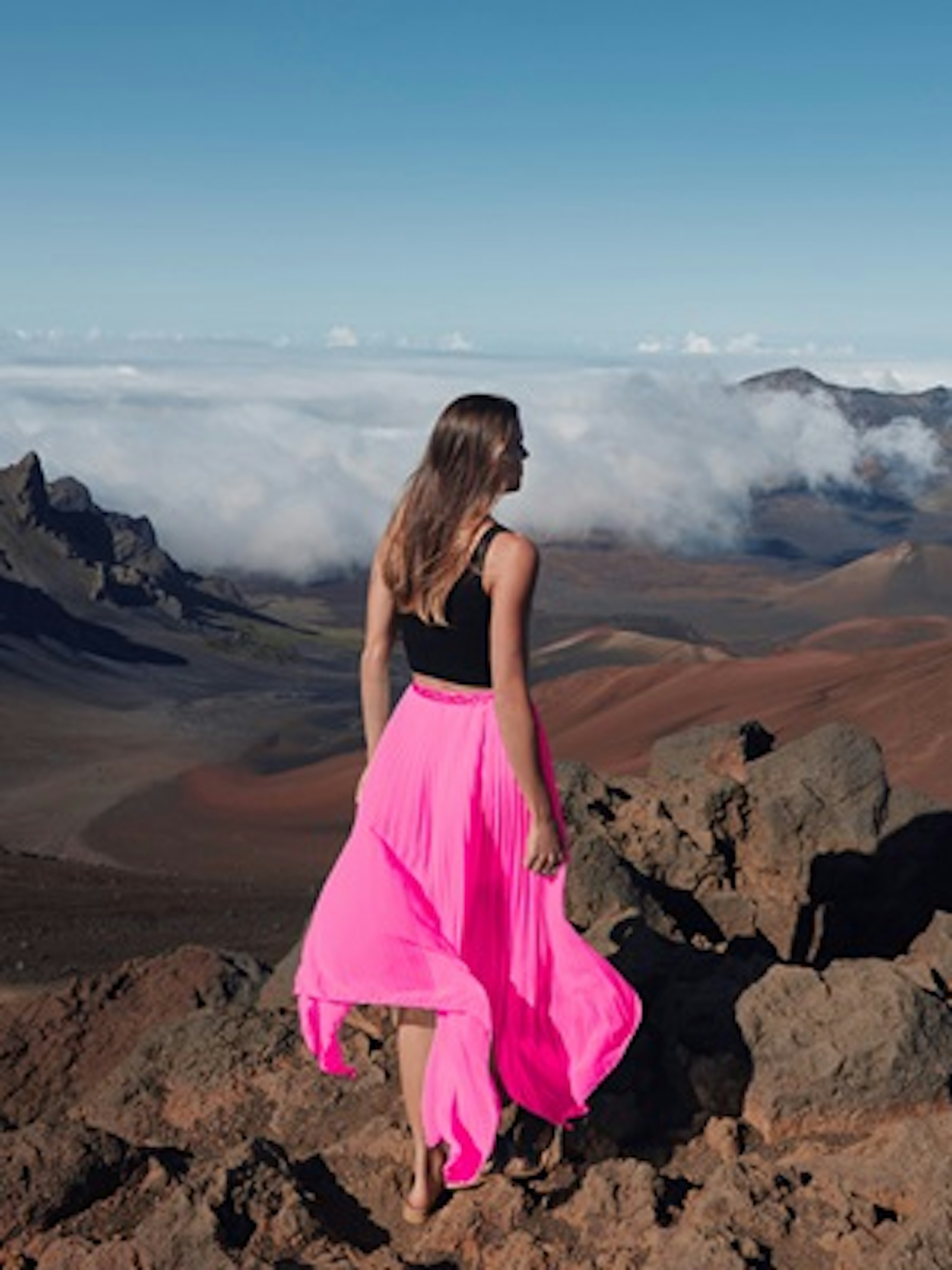 Une femme portant un haut noir et une longue jupe rose vif se tient sur un sommet de montagne rocheux, contemplant un vaste paysage montagneux et spectaculaire. Le ciel est dégagé, avec quelques nuages au-dessus des montagnes.