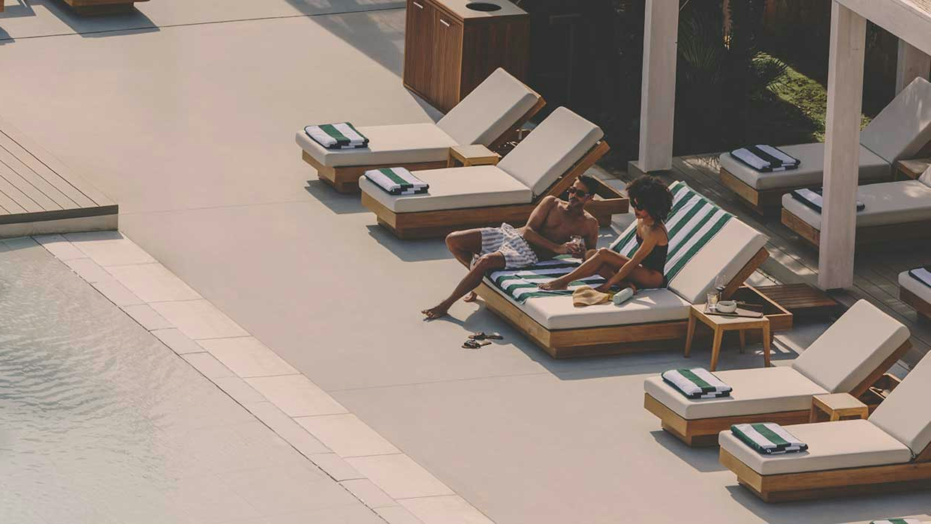 Aerial view of a luxurious outdoor pool area with several lounge chairs. Two people relax on a striped lounge chair by the pool.