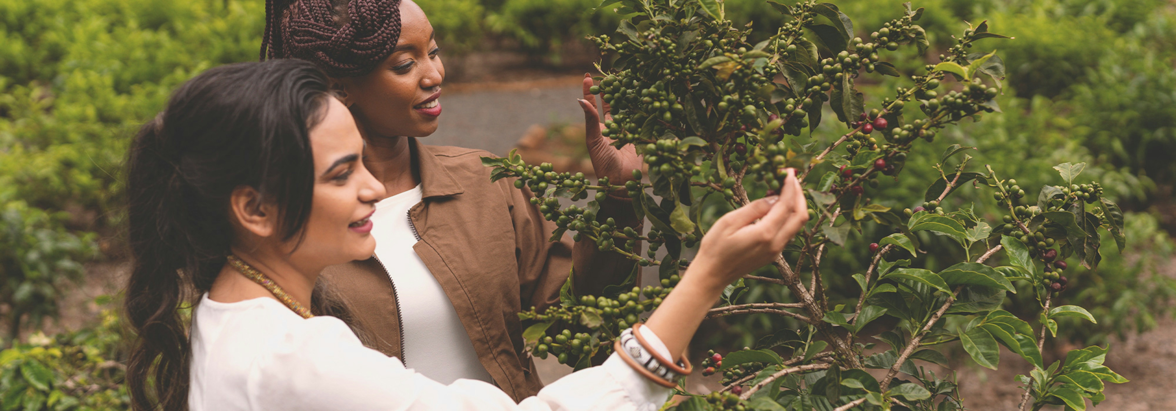 Deux femmes en extérieur, l'une aux cheveux tressés portant une veste marron, l'autre aux cheveux lisses portant un haut clair, toutes deux regardant la verdure.