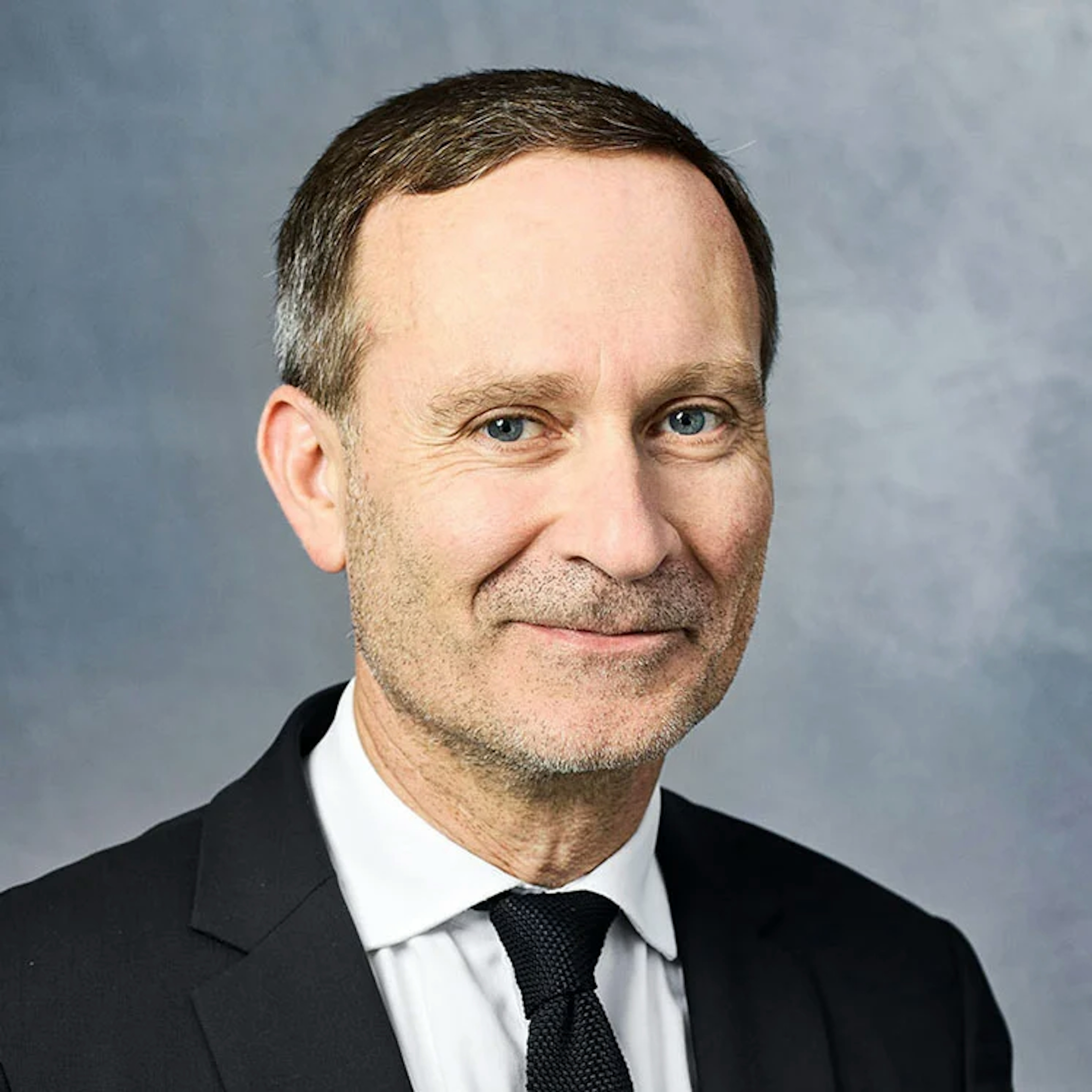 Portrait of Pierre Boisselier, a man with short brown hair and blue eyes, wearing a black suit, white shirt, and black tie, smiling.