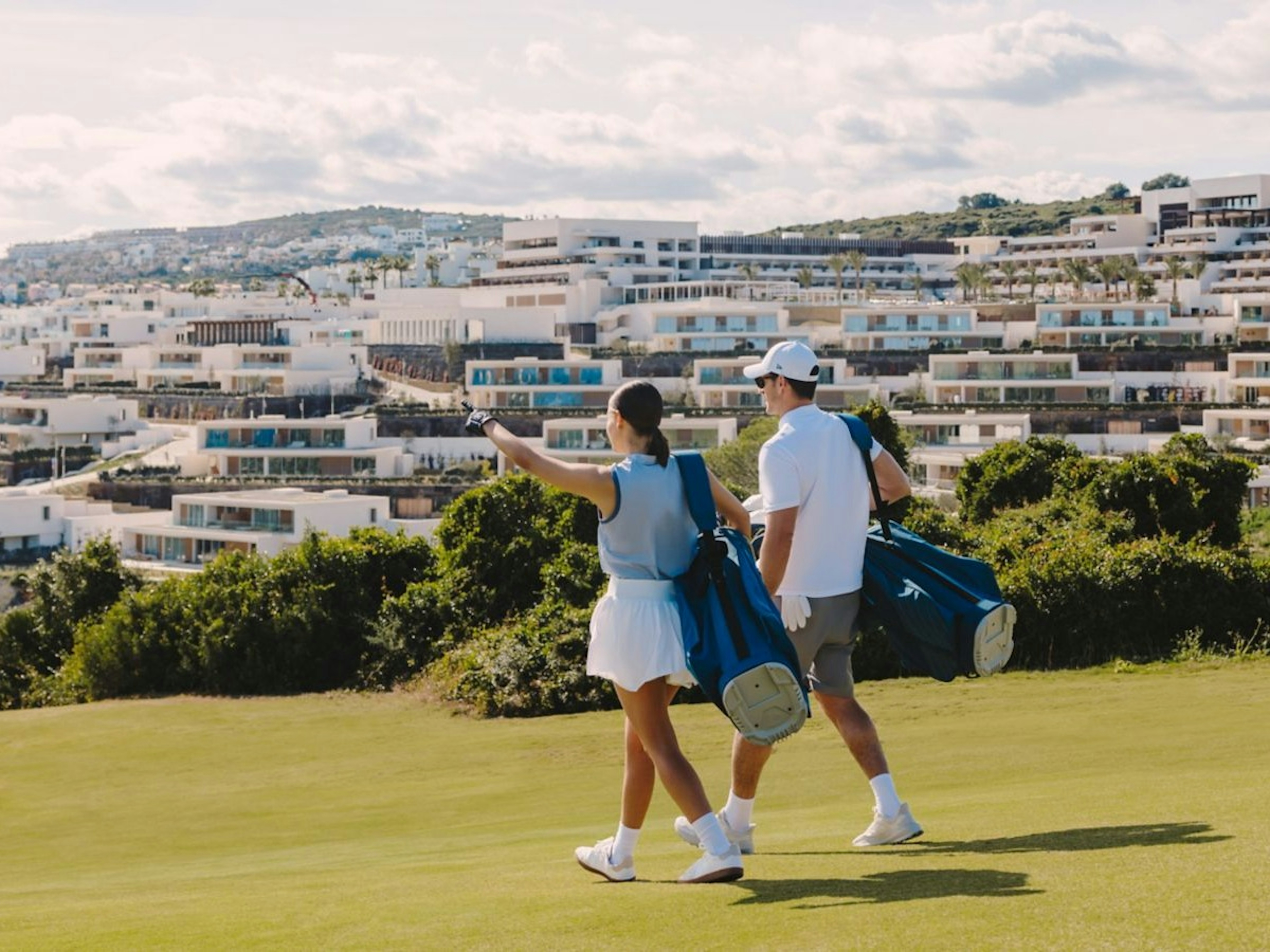 Un couple marchant sur l’herbe verte avec du matériel de golf, souriant en désignant un endroit sur le parcours où ils prévoient de jouer.