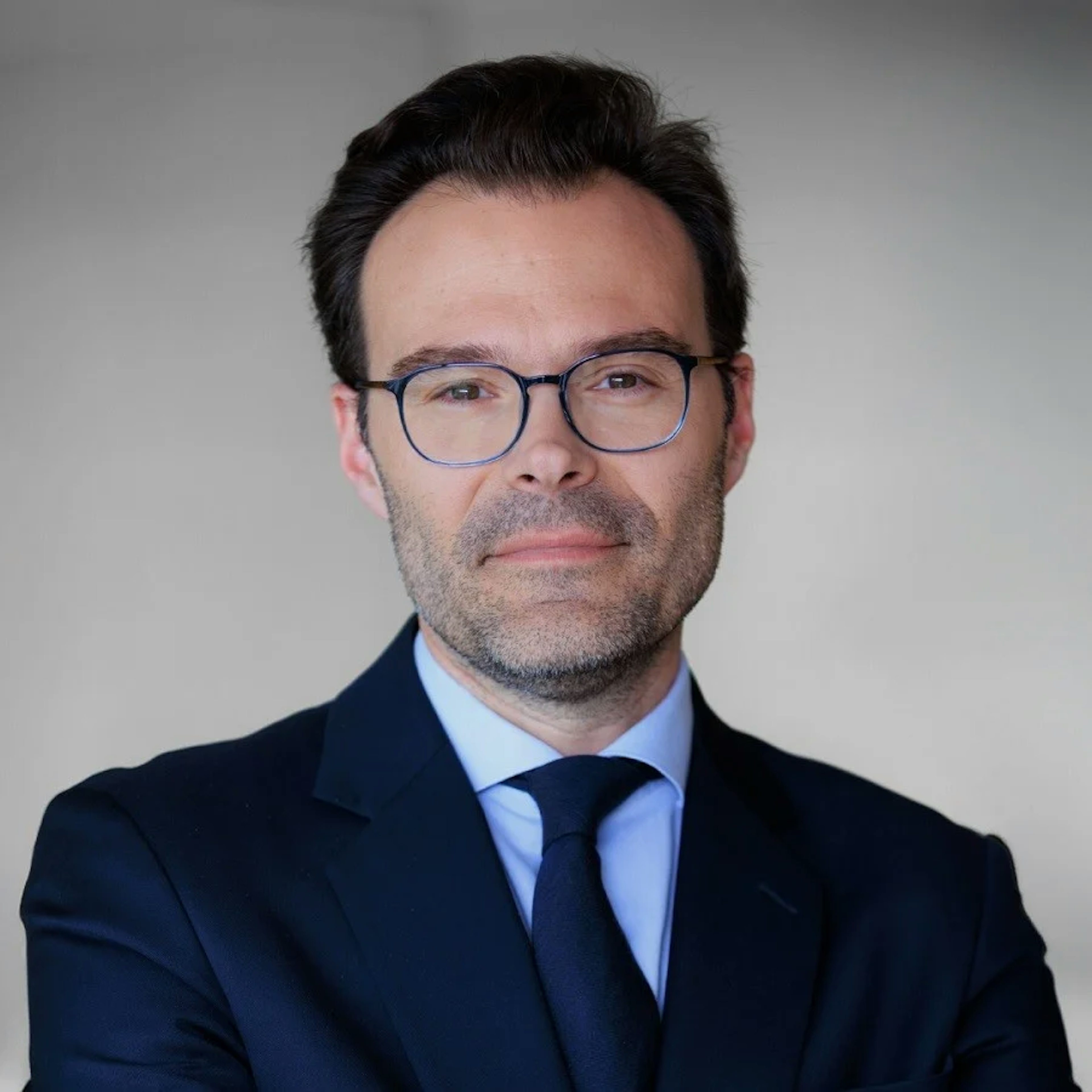 Portrait of Pierre Deburaux, a man with dark hair, glasses, wearing a dark suit and blue tie, looking at the viewer with a neutral expression.